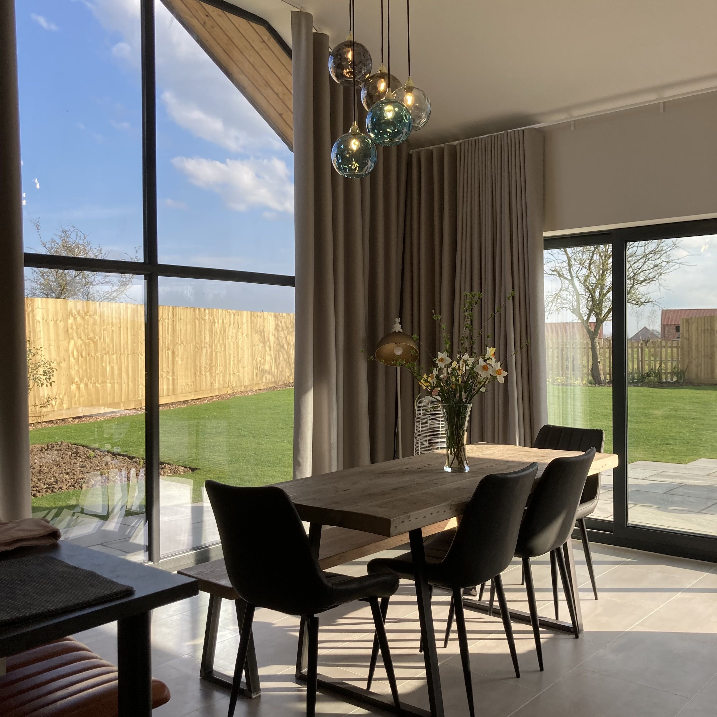 A modern dining area with a wooden table and black chairs, large windows, and a view of a backyard.