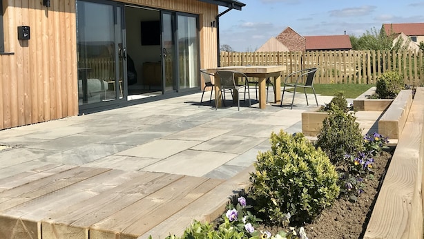 Une maison en bois avec une porte d’entrée en verre, une terrasse avec une table et des chaises, et un jardin fleuri.