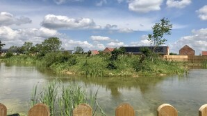 Une vue paisible sur un lac, avec une clôture en bois au premier plan, une étendue d’eau calme et, au loin, des bâtiments aux toits rouges sous un ciel partiellement nuageux.