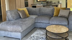 A modern living room with a grey sectional sofa, a round wooden coffee table, and a kitchen in the background. - Water Lily Cottage, Ashlin Farm Barns (Lincoln)