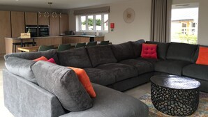 A modern living room with a grey sectional sofa, a round black coffee table, and a dining area with wooden cabinets and a kitchen in the background. - Chestnut House, Ashlin Farm Barns (Lincoln)