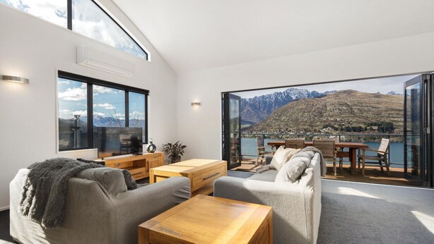 Una sala de estar moderna con un ventanal grande, un sofá, una mesa de centro y un comedor con vistas a montañas y un lago.