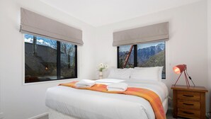 Un dormitorio con cama, dos ventanas con vistas a la montaña, una lámpara de noche y una mesita de noche.