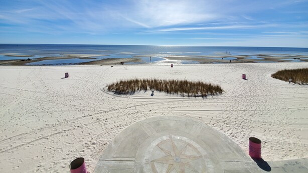 Ein Strand mit blauem Himmel, Sandfläche mit Fußabdrücken und einer runden Steinarchitektur mit Kompassmuster.