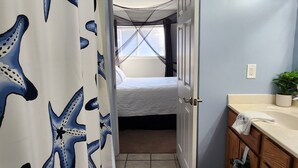 Ein Badezimmer mit Duschvorhang mit Sternfisch-Muster, einem Bett mit weißer Matratze, einem Holzschrank mit Handtuch und einer Topfpflanze auf der Ablagefläche.