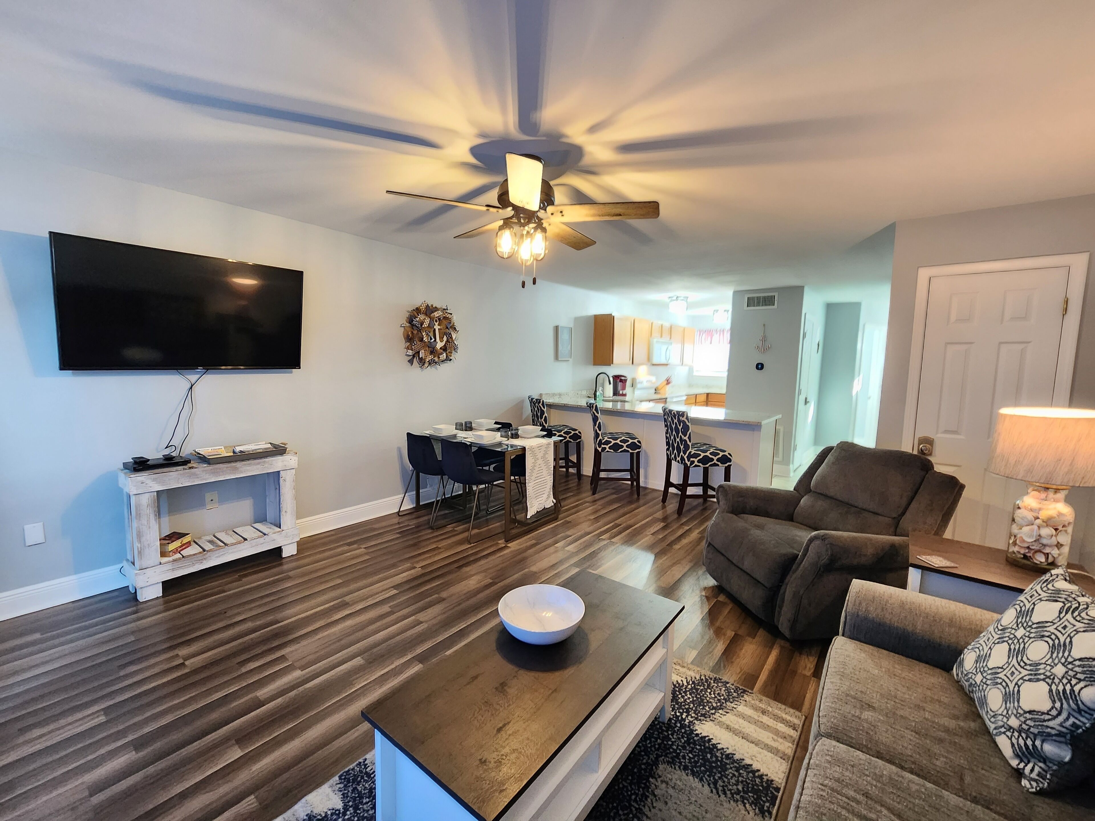 A modern living room with a flat-screen TV, a ceiling fan, and a dining area with a kitchen in the background.