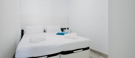 A small, modern bedroom with a single bed, white bedding, and a nightstand with a lamp.