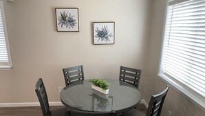 En matplats med ett runt bord i glas, omgivet av fyra stolar. På väggen hänger två inramade botaniska illustrationer, och det finns ett fönster med persienner.