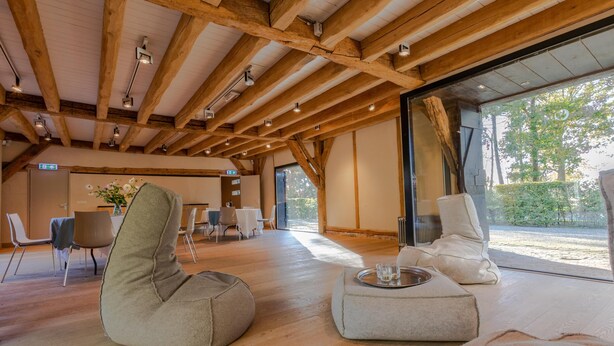 Een ruime kamer met houten balken, een groot raam en moderne meubels, waaronder een relax fauteuil en een comfortabele zitting.