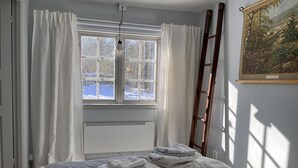 Un dormitorio con cama, radiador, ventana con cortinas, una escalera y un cuadro en la pared.