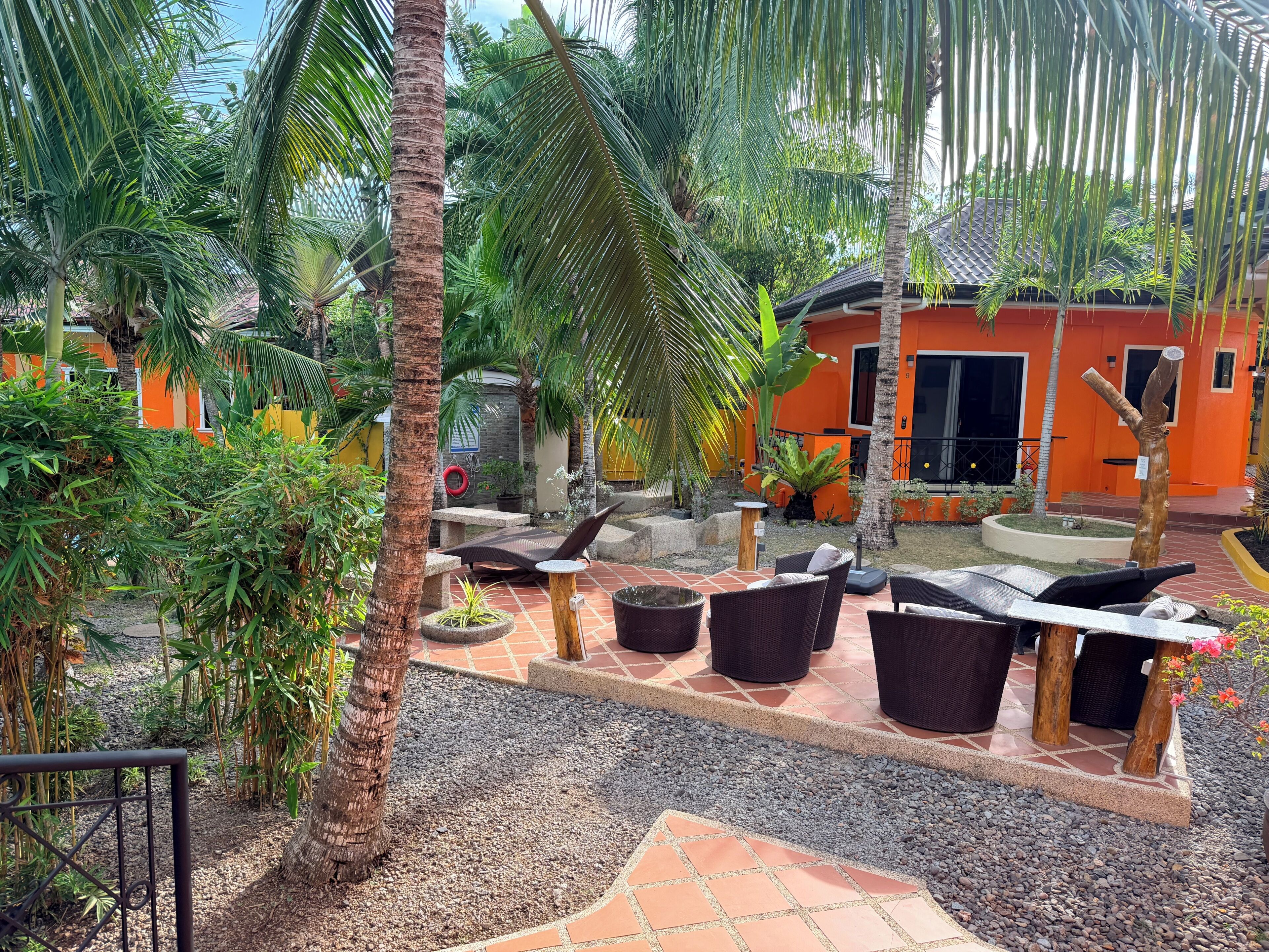 A tropical outdoor seating area with lounge chairs and tables, surrounded by palm trees and potted plants.
