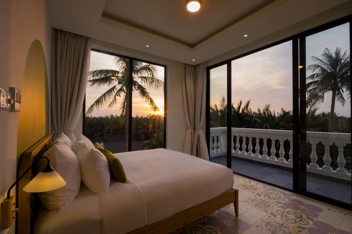 A bedroom with a large bed, a nightstand, a lamp, and a view of palm trees and a balcony.