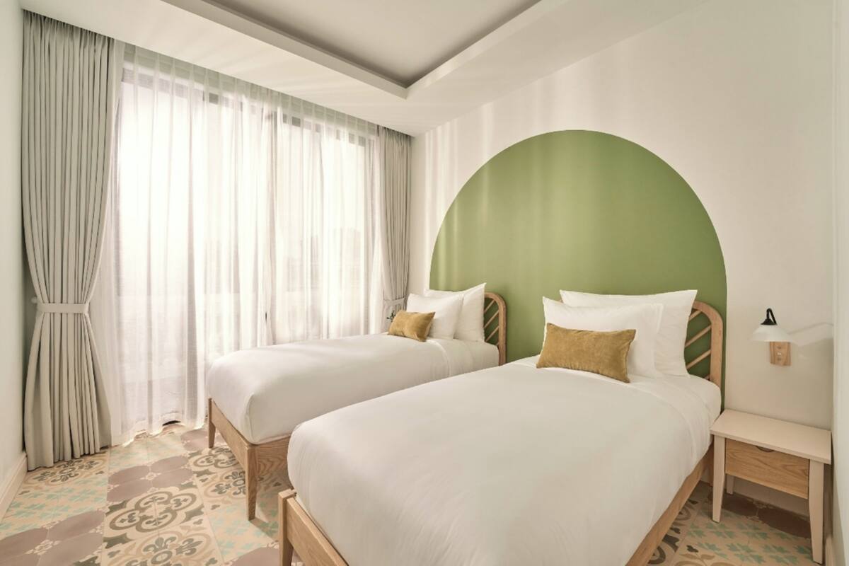 A hotel room with two beds, a large window with sheer curtains, a green accent wall, and a patterned floor.
