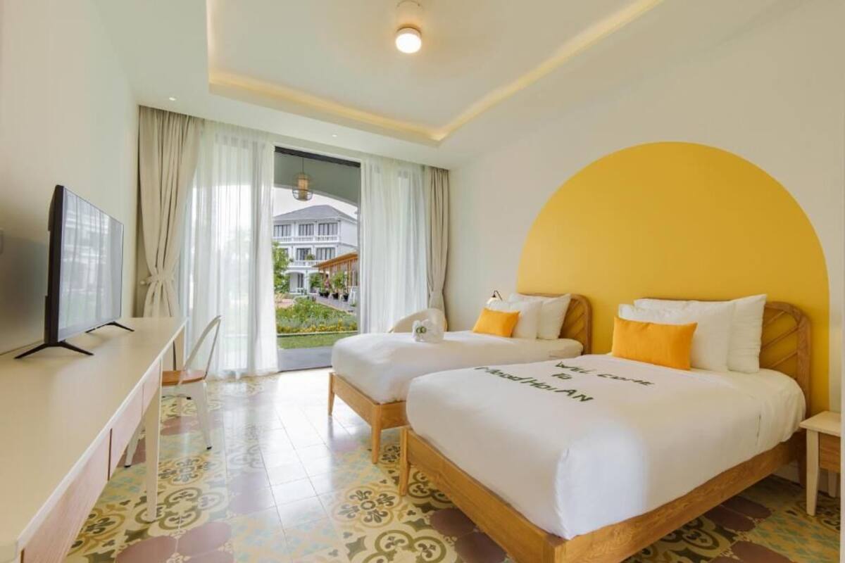 A hotel room with two beds, a TV, a dining table, and a view of a building outside.