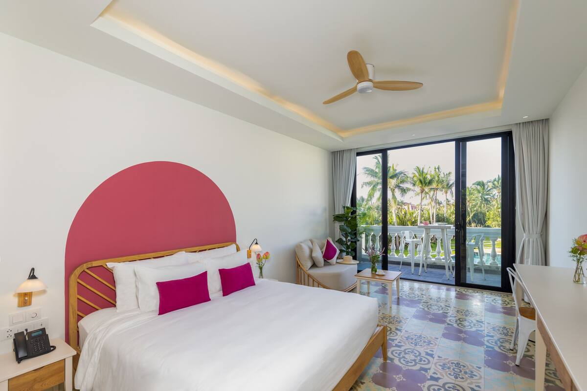 A hotel room with a bed, a desk, a chair, a balcony with a view of palm trees, and a ceiling fan.
