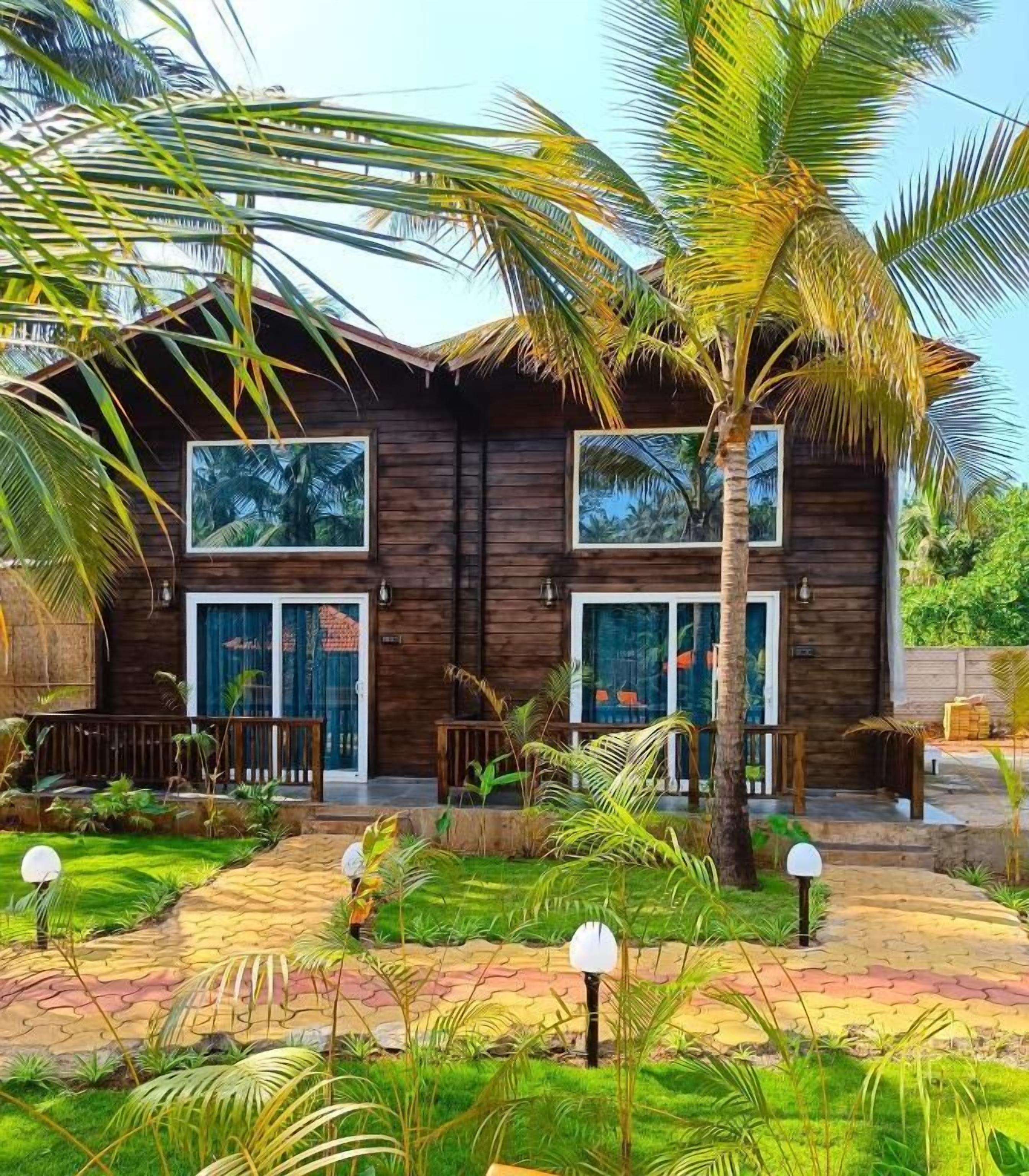 A two-story wooden house with large windows and a well-maintained garden with palm trees and other tropical plants.
