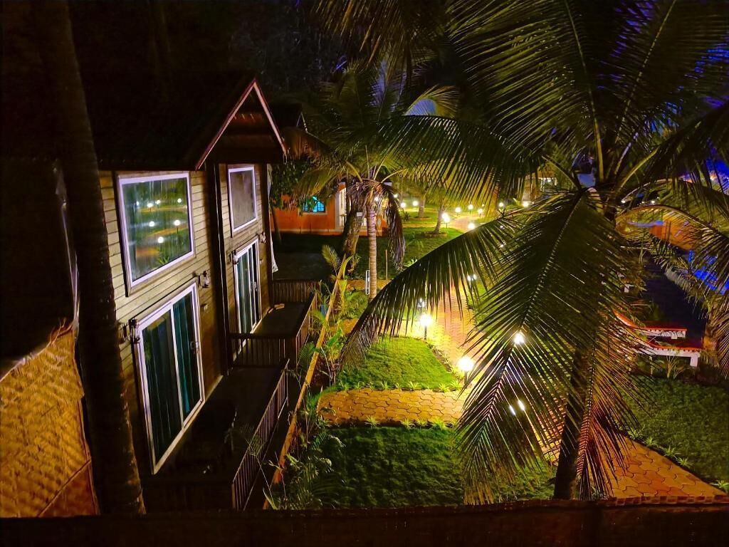A well-lit, two-story building with a balcony, surrounded by lush greenery and palm trees.
