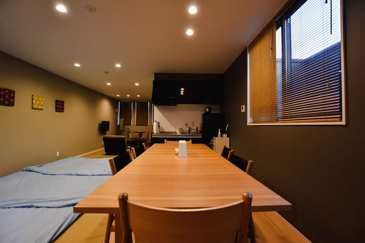 A room with a long wooden table, several chairs, a kitchenette, and a window with blinds.