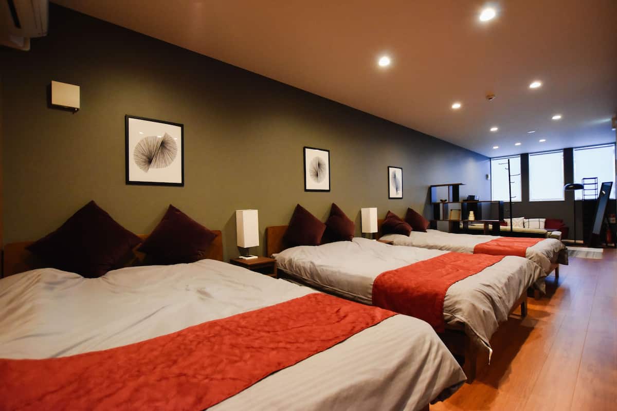 A hotel room with four beds arranged in a row, each with a red blanket. There are framed artworks on the walls and a large window providing natural light.