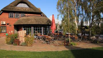 A traditional red building with a thatched roof, outdoor seating area with tables and chairs, and a garden with plants and trees.