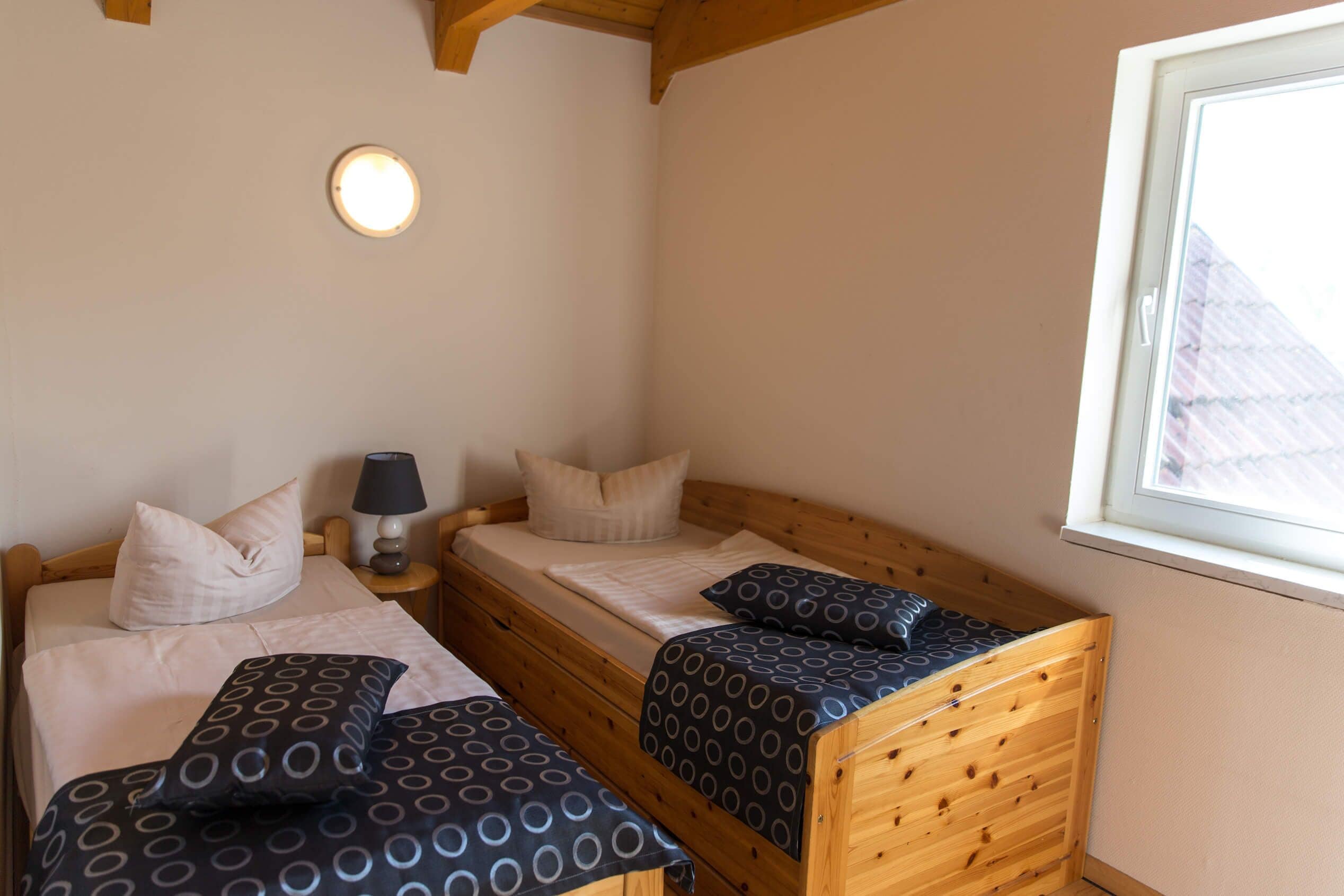 Ein Schlafzimmer mit zwei Holzbettgestellen übereinander, einem Nachttisch mit Lampe und einem Fenster mit Blick auf ein Gebäude.