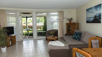 Sala de estar con sofá, televisor, mesa con un florero y vista a un jardín por un ventanal corredero.