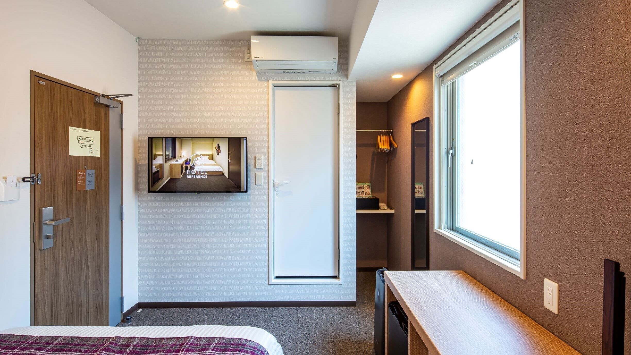 A hotel room with a bed, a TV mounted on the wall, a wardrobe, a window, and an air conditioning unit.
