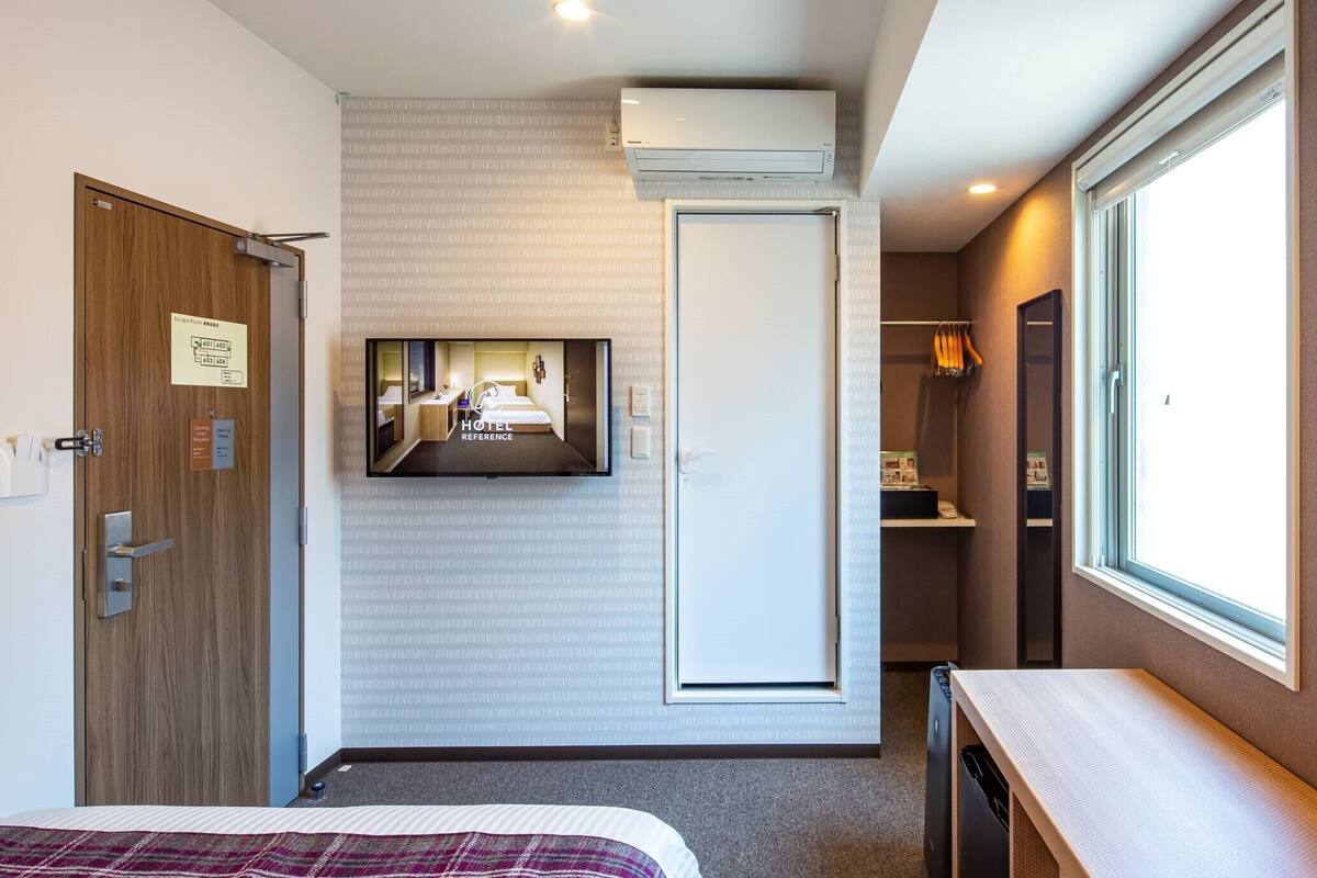 A hotel room with a bed, a TV mounted on the wall, a wardrobe, a window, and an air conditioning unit.