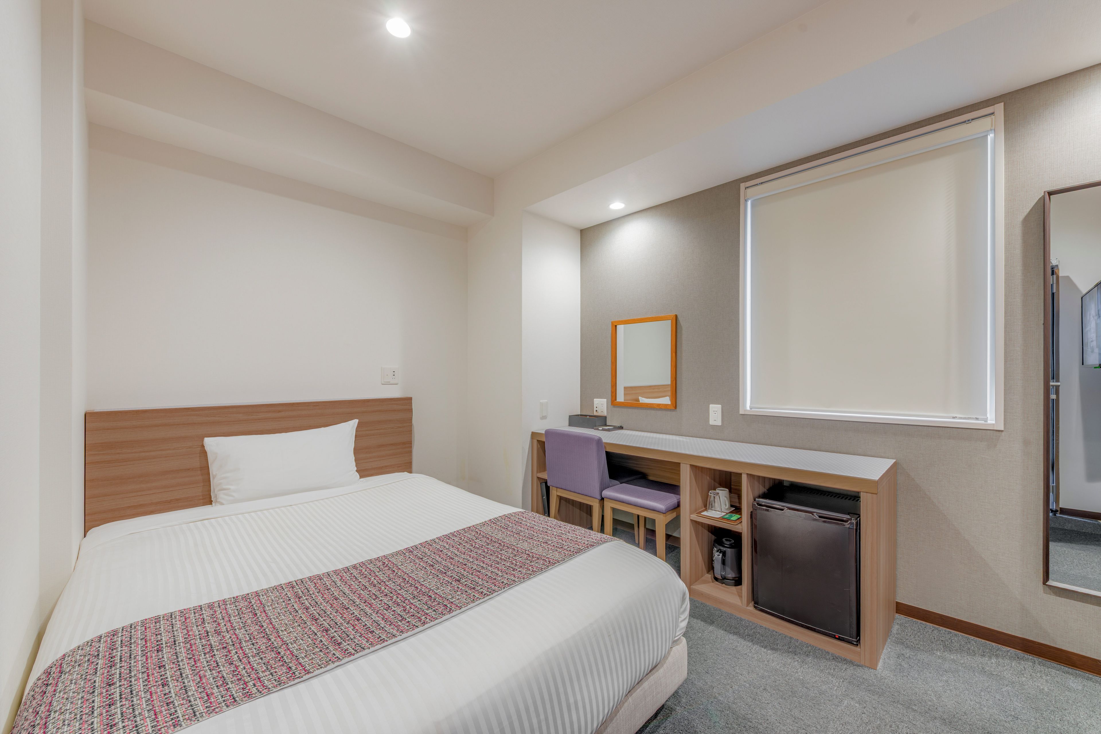 A hotel room with a bed, a desk, a chair, a small refrigerator, and a mirror.