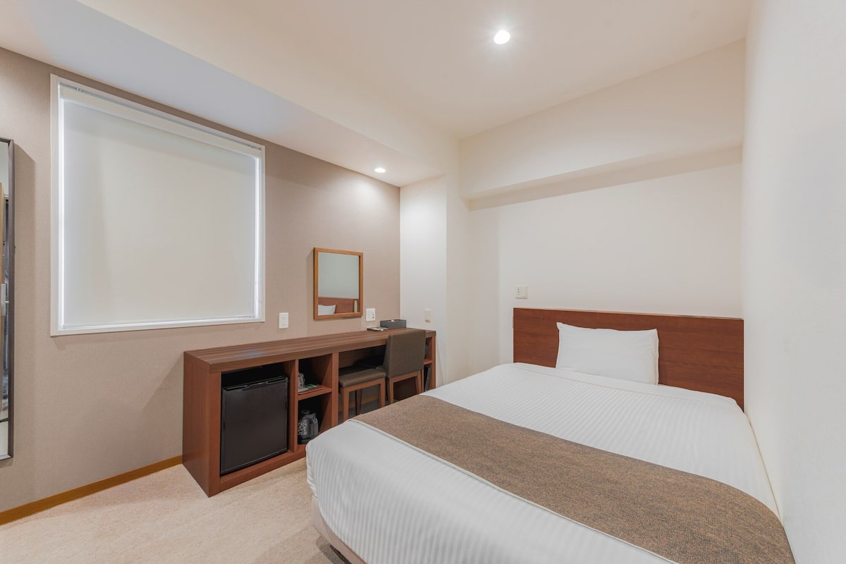 A hotel room with a bed, a desk, a chair, a small cabinet, a mirror, and a window.