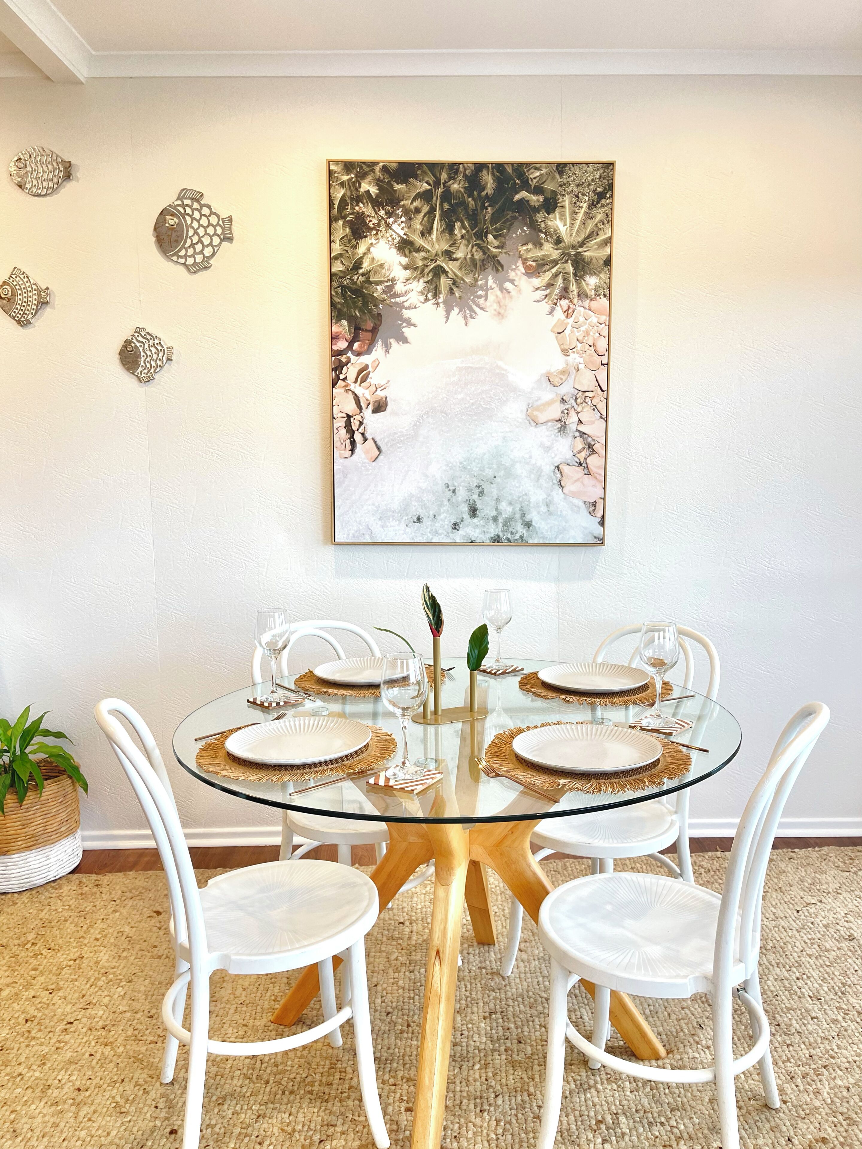 Un coin repas avec une table en verre, des chaises blanches et un tableau aux motifs tropicaux accroché au mur.