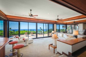 A spacious living area with a large sliding glass door leading to a balcony with a view of the ocean. There are multiple seating areas, including a sofa and two chairs, and a small bar area with stools. - Villas at Mauna Kea 14 Two Bedroom (Kamuela)