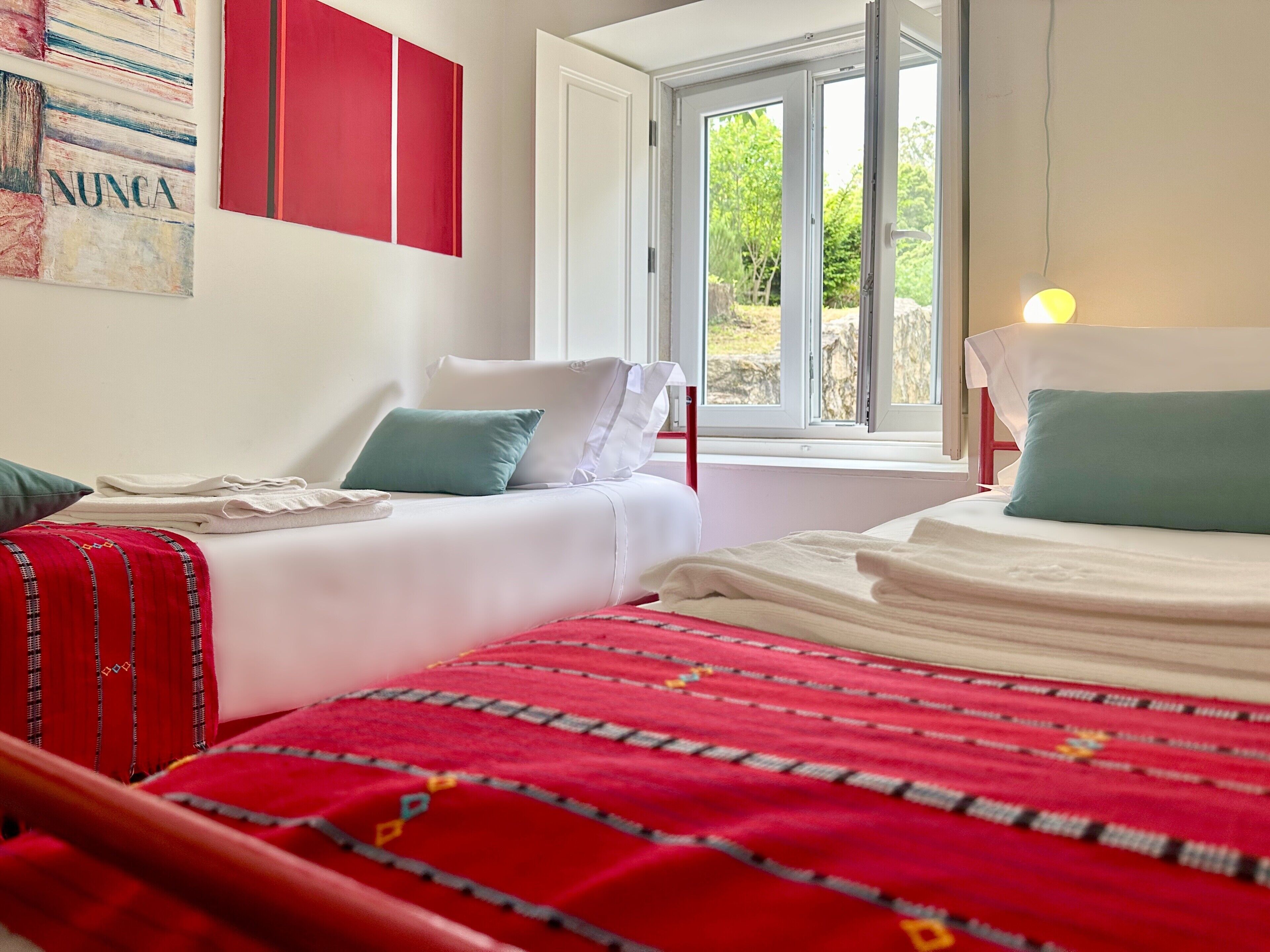 Un dormitorio con dos camas, una ventana con vistas a la vegetación y un adorno rojo y blanco en la pared.