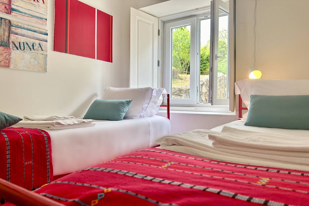 Une chambre avec deux lits, une fenêtre donnant sur un paysage verdoyant et une décoration murale aux couleurs rouge et blanche.