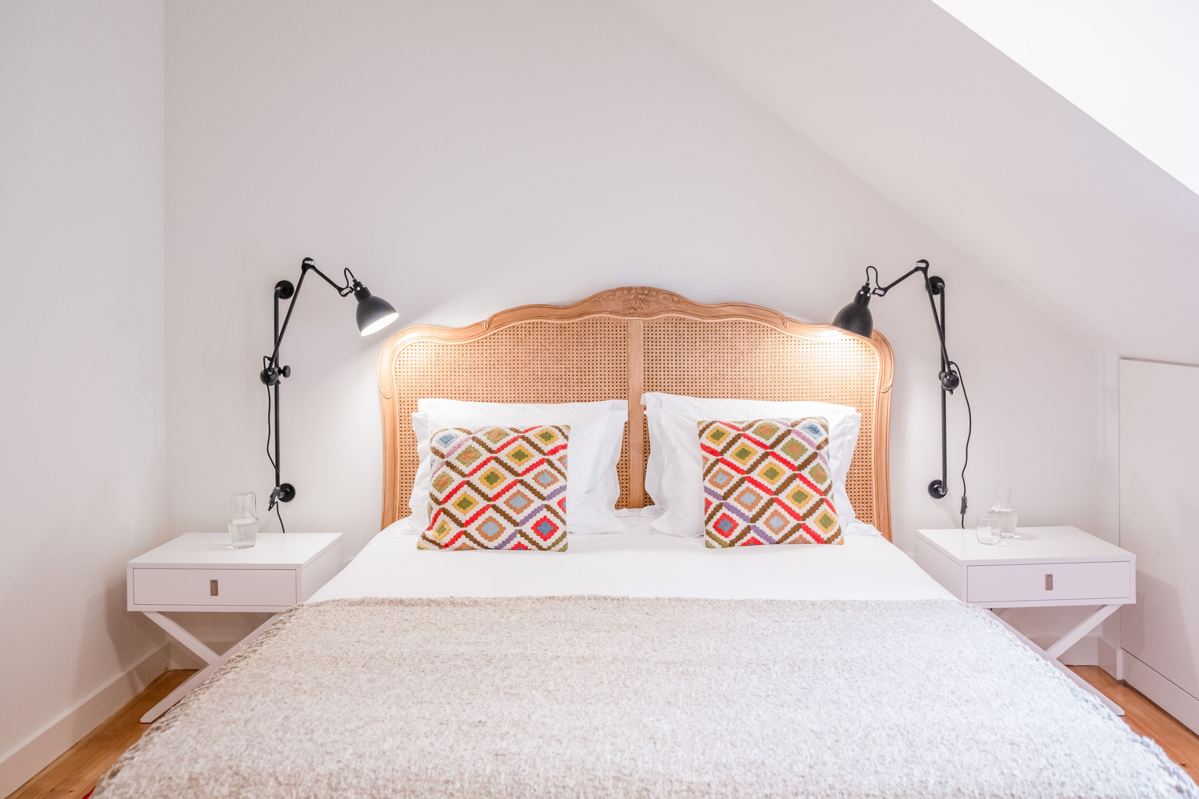 A neatly arranged bedroom with a rattan headboard, two bedside tables with lamps, and patterned pillows.