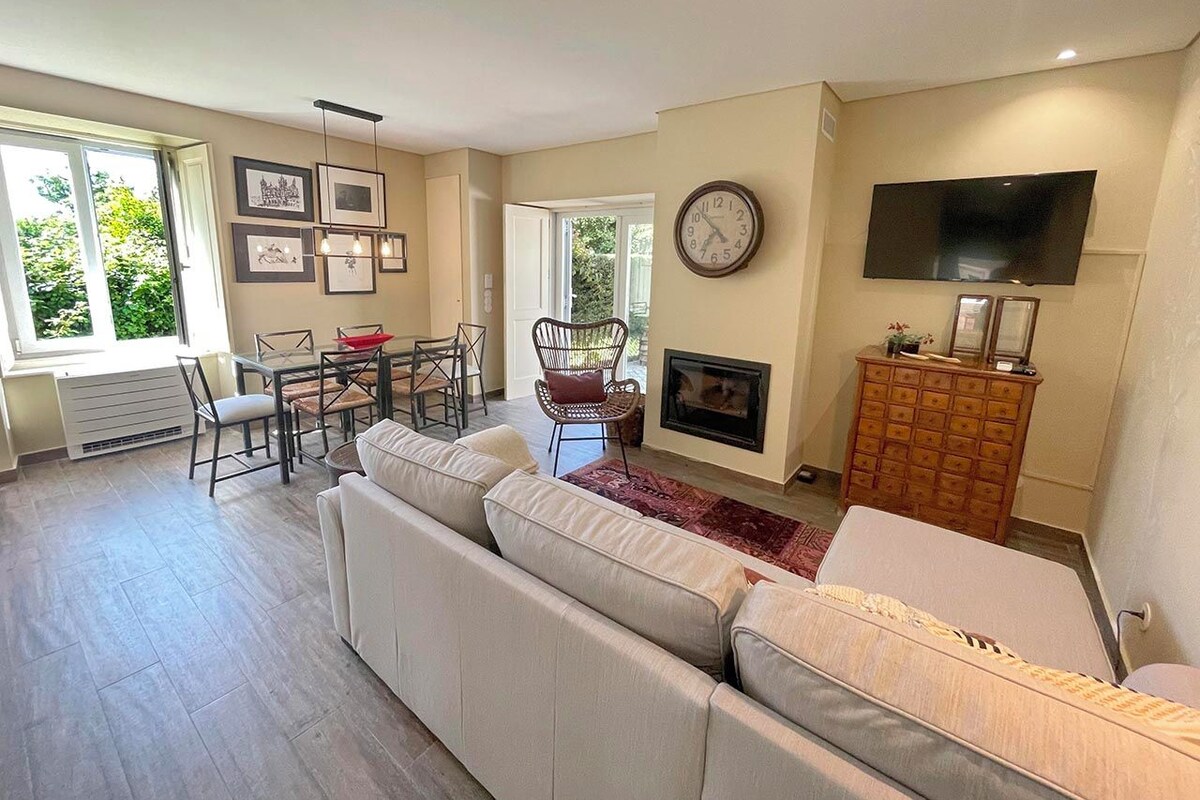 A spacious living room with a large sofa, a dining table with chairs, a fireplace, a television, and framed pictures on the wall.