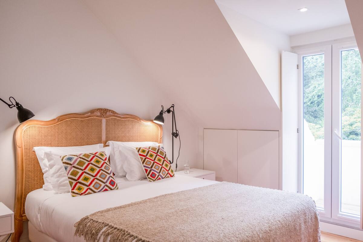 Une chambre avec un plafond en pente, un lit doté d’une tête de lit en osier, deux lampes de chevet et une grande fenêtre donnant sur un paysage verdoyant.