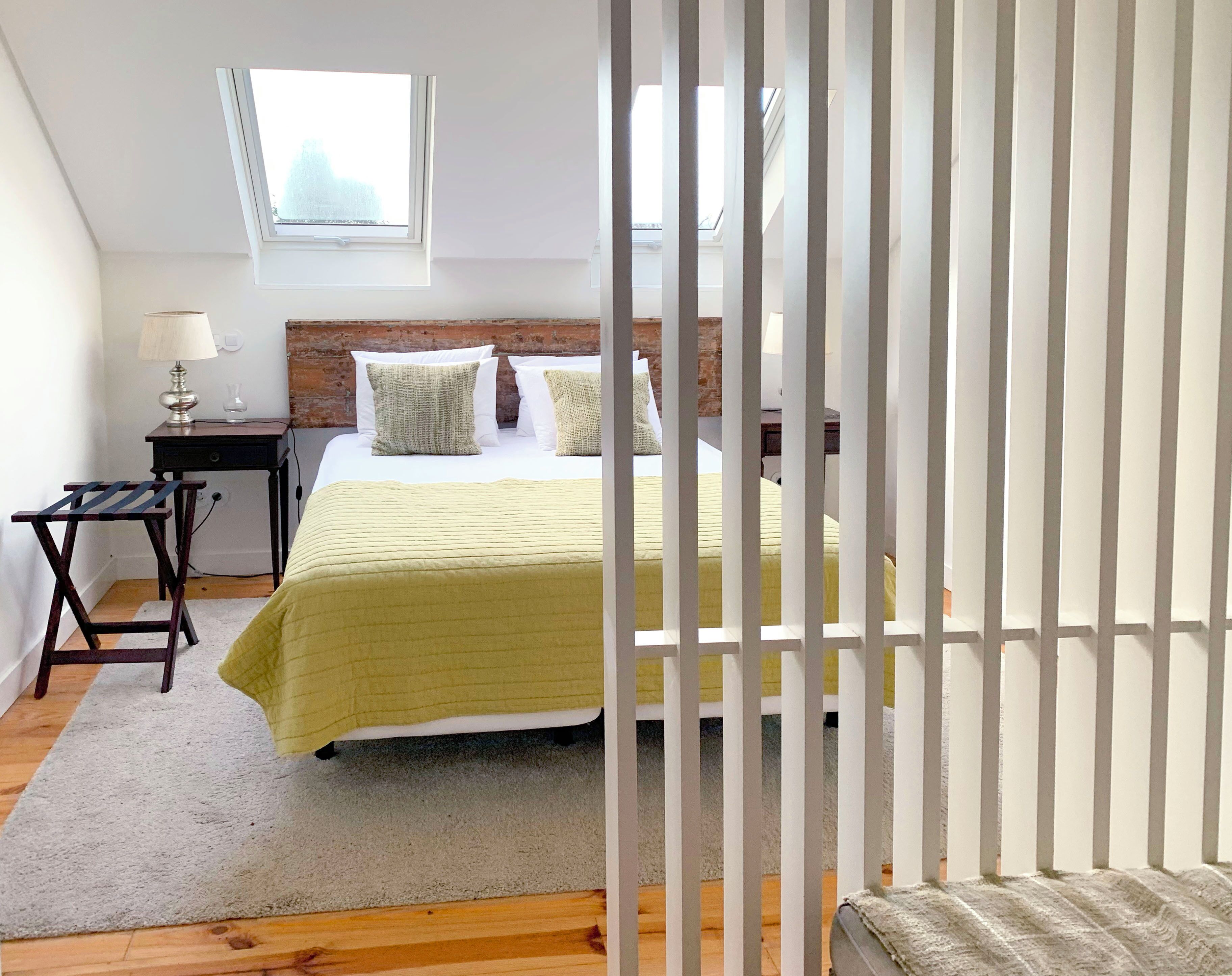Un dormitorio con cama, mesita de noche, lámpara, ventana y vista al cielo.