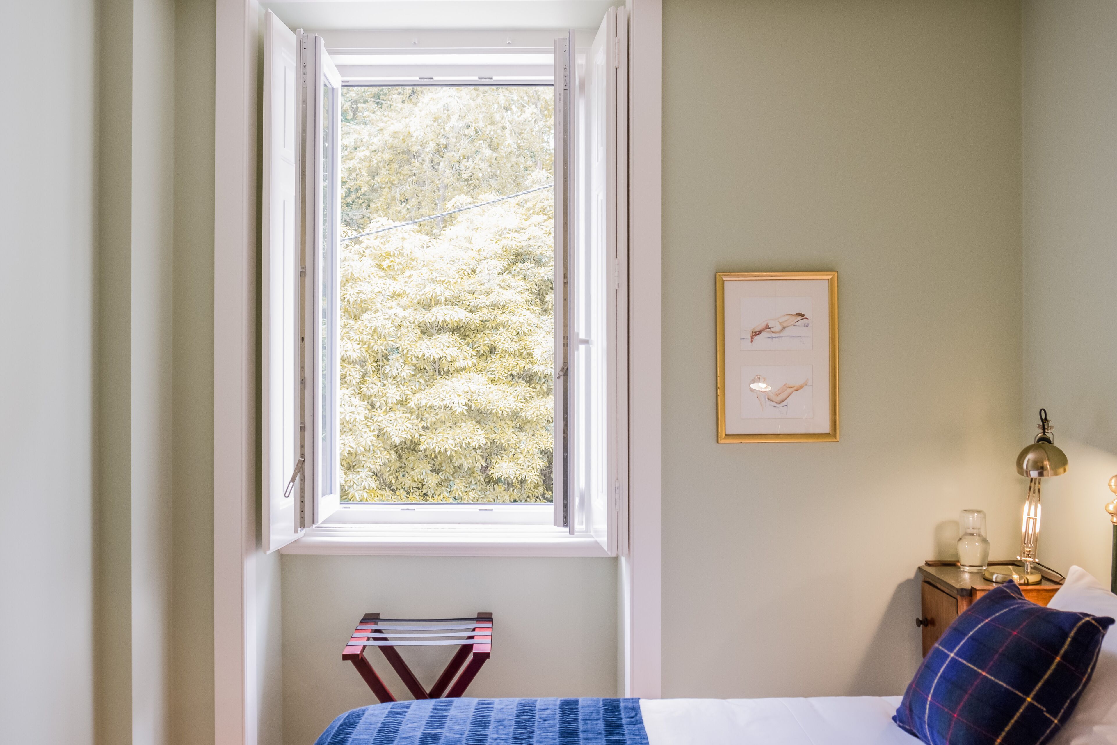 Une chambre avec une fenêtre, une table de chevet avec une lampe et un tableau encadré au mur.