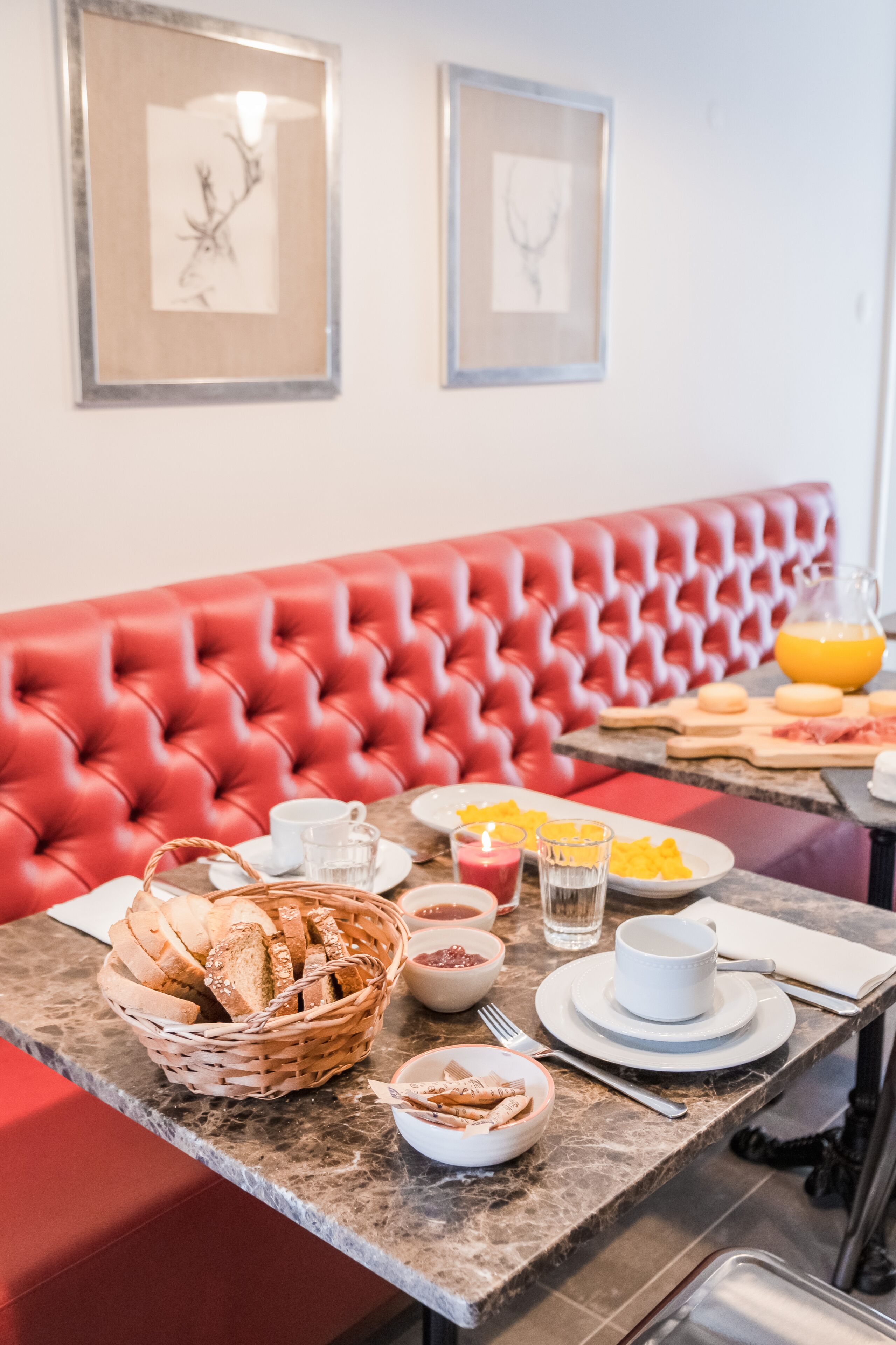 Una mesa de desayuno con pan, pastelería y bebidas, ubicada en un asiento tapizado de rojo con cuadros en la pared.