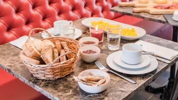 Una mesa de desayuno con pan, pastelería y bebidas, ubicada en un asiento tapizado de rojo con cuadros en la pared.