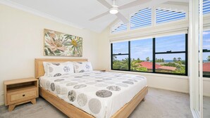 A bedroom with a large bed, a nightstand, a ceiling fan, a window with a view of the sea, and a floral painting on the wall. - HAVENVIEW TERRACES #2 | Close to beach, shops and cafes | Accom Property (TERRIGAL)