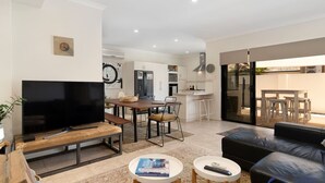 A modern living room with a flat-screen TV, a wooden bench, a dining table with chairs, and a black leather sofa. - Summer Breeze at Avalon Beach (Wannanup)