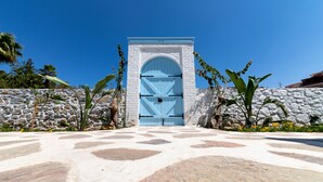 A stone wall with a blue gate, surrounded by tropical plants and a clear blue sky. - Louise'in Konağı (Datça)