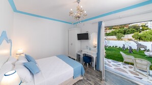 A bedroom with a bed, a desk, a TV, and a view of a garden with statues. - Louise'in Konağı (Datça)