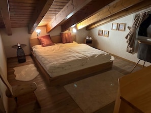 A cozy bedroom with a large bed, wooden beams, a small bedside table, a nightstand, and a window with curtains.