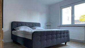 Een slaapkamer met een donkergrijs gestoffeerd bed, wit beddengoed en een groot raam dat natuurlijk licht binnenlaat.