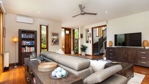 A spacious living room with a large grey sofa, a wooden coffee table, and a television on a cabinet. There is a bookshelf filled with books and a potted plant. - Pet Friendly - Bayside Oasis in Highett (Highett)