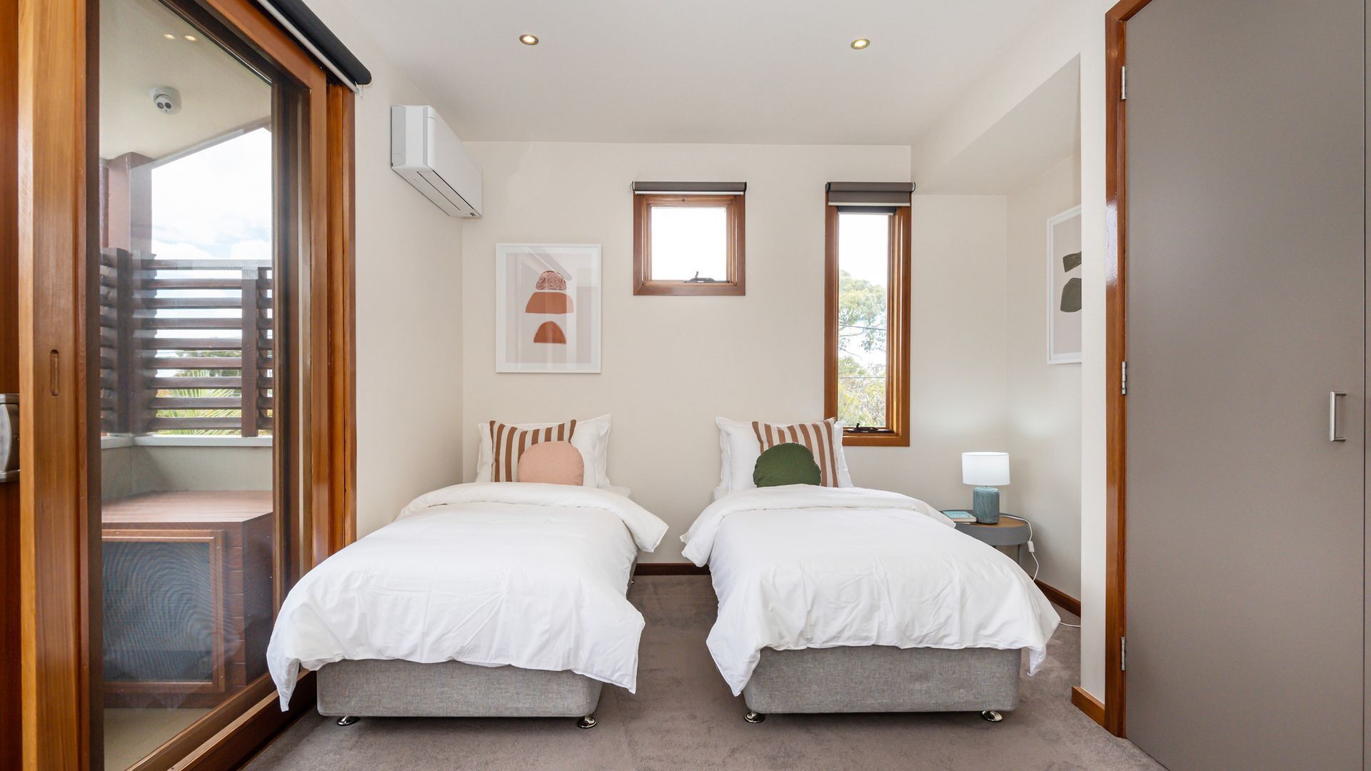 A bedroom with two beds, a window, an air conditioning unit, and a small table with a lamp.