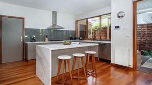 A modern kitchen with a central island, wooden stools, a stainless steel range hood, and a view of the outdoors. - Pet Friendly - Bayside Oasis in Highett (Highett)